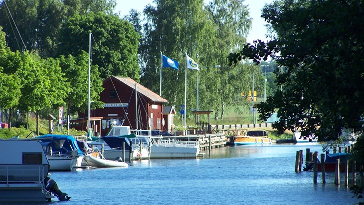En bild över Torshälla gästhamn sommartid. Vatten, båtar, en röd träbyggnad och och träd.