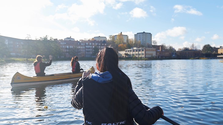 Bild tagen bakifrån på en flicka som paddlar kanot i Eskilstuna ån. Ytterligare en kanot med två flickor till vänster. Eskilstuna stad i bakgrunden.